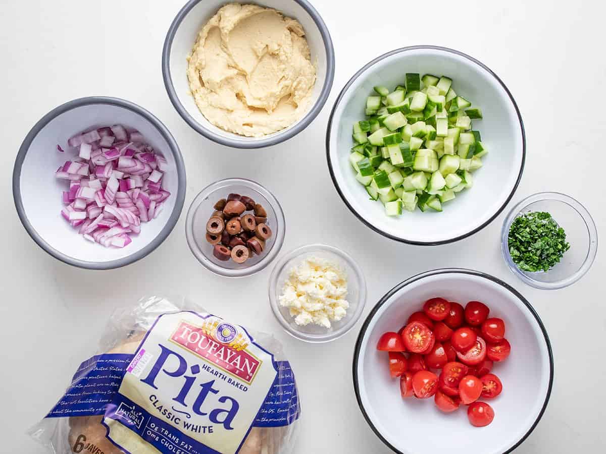 Overhead shot of ingredients, including pitas, diced onions, sliced kalamata olives, crumbled feta, sliced tomatoes, and chopped parsley.