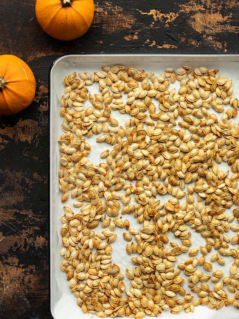 roasted pumpkin seeds on a baking sheet with pumpkins on the side