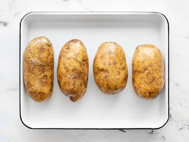 Four prepped russet potatoes on a small white baking sheet