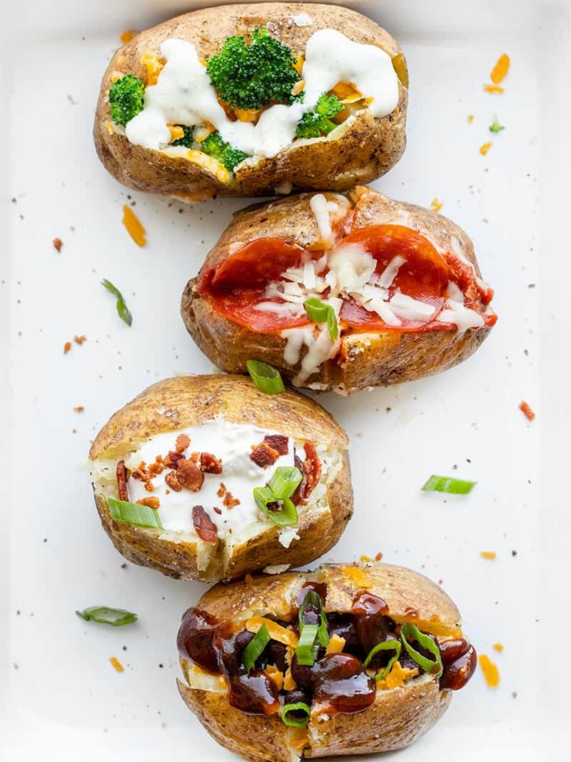 Four baked potatoes lined up on a baking sheet, each topped with different ingredients