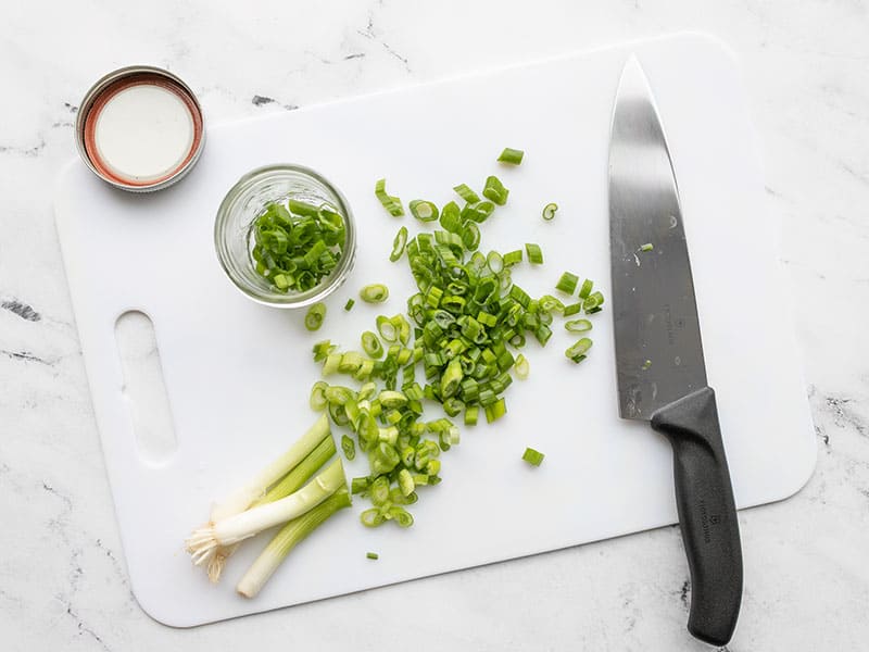 Sliced green onion on a cutting board with a chef's knife and mason jar on the side