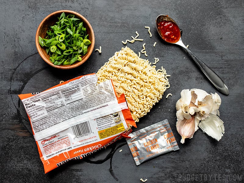 An opened package of instant ramen with other ingredients around it, including green onions, chile paste, and garlic.