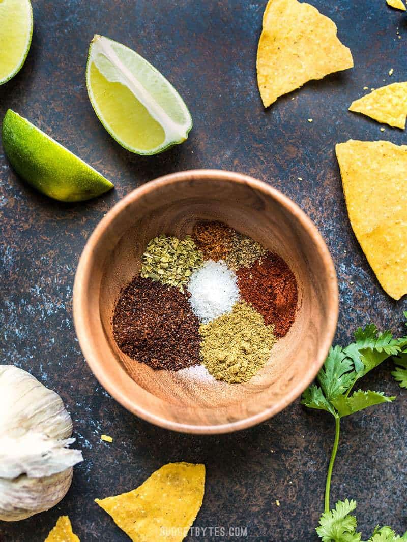 Individual ingredients for homemade taco seasoning in a small wooden bowl, surrounded by chips, limes, cilantro, and garlic. 
