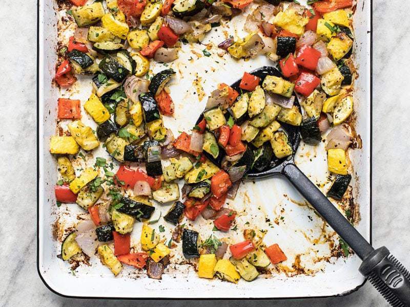 Roasted summer vegetables on a sheet pan being scooped up by a spatula. 