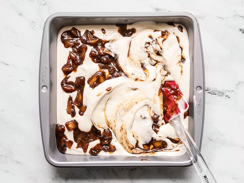 Whipped ice cream mixture and balsamic peaches in a baking dish being swirled with a spatula.