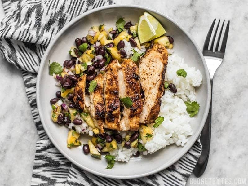 Sliced Jerk Chicken with Pineapple Black Bean Salsa on a bed of rice on a plate with a black and white napkin.