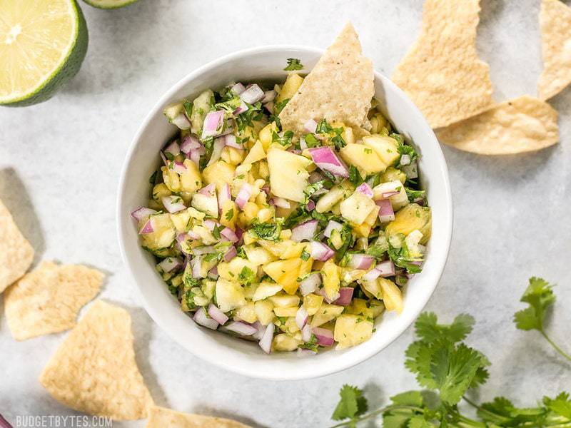 A bowl of easy pineapple salsa with chips, lime, and cilantro around the bowl. 