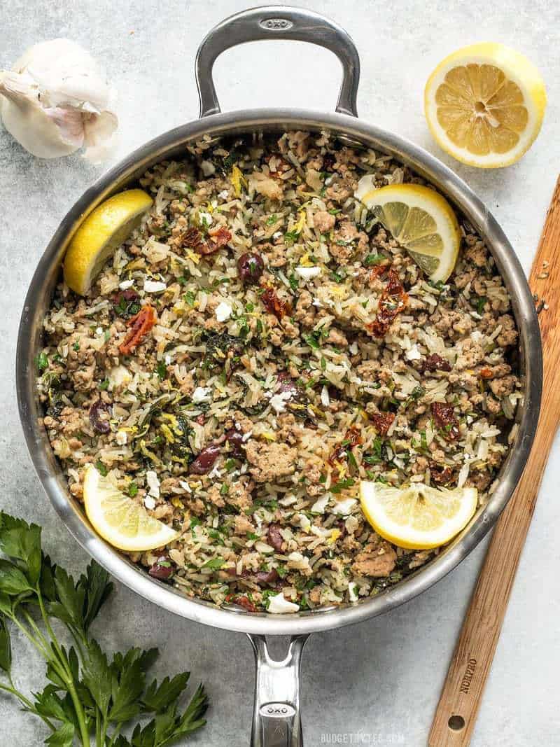 A large skillet full of Greek Turkey and Rice Skillet with lemon wedges, garlic, and parsley on the sides