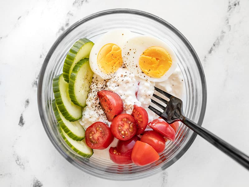 A glass bowl with cottage cheese, hard boiled egg, tomatoes, cucumber, and pepper.