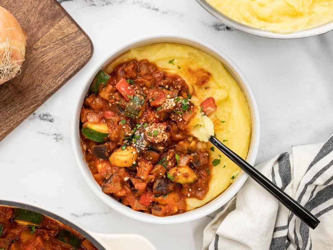 A bowl of summer vegetables in red sauce over polenta with a fork in the center.