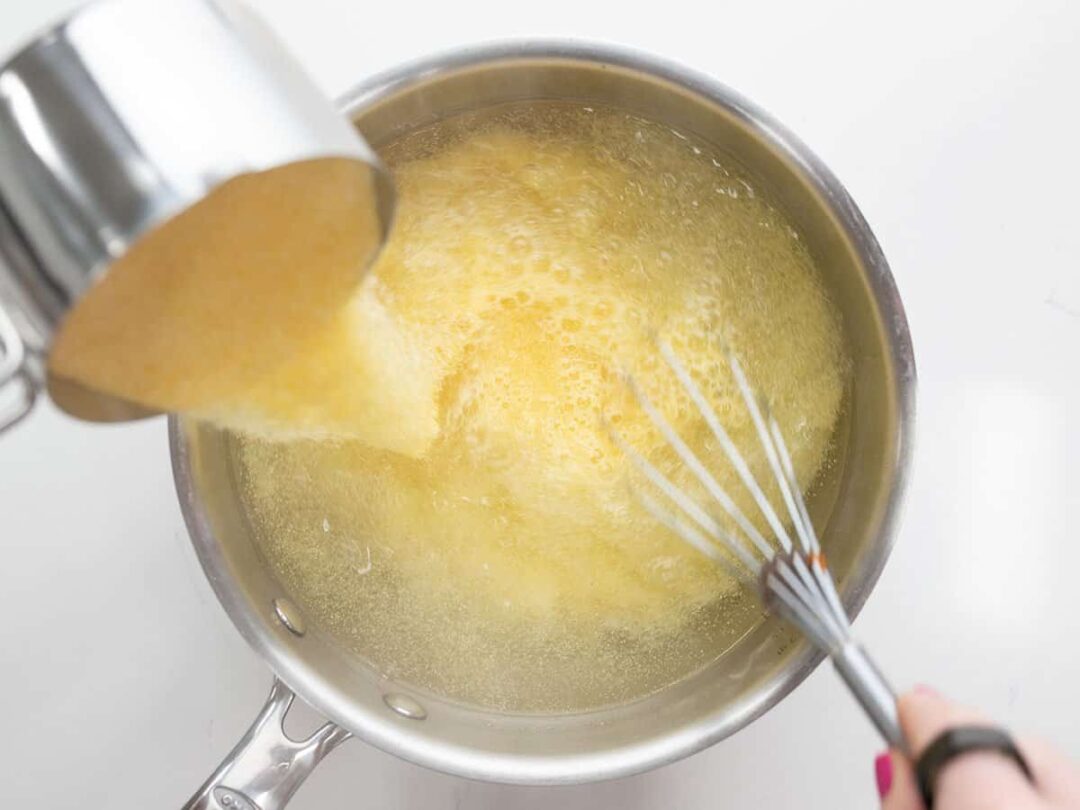 Cornmeal being poured into a pot of boiling water with a whisk.