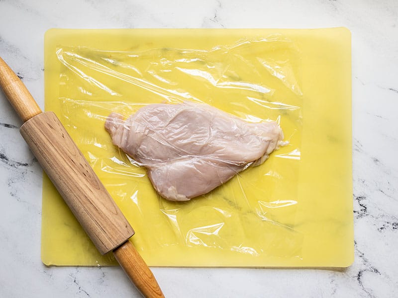 Pounded chicken on a cutting board with rolling pin on the side.