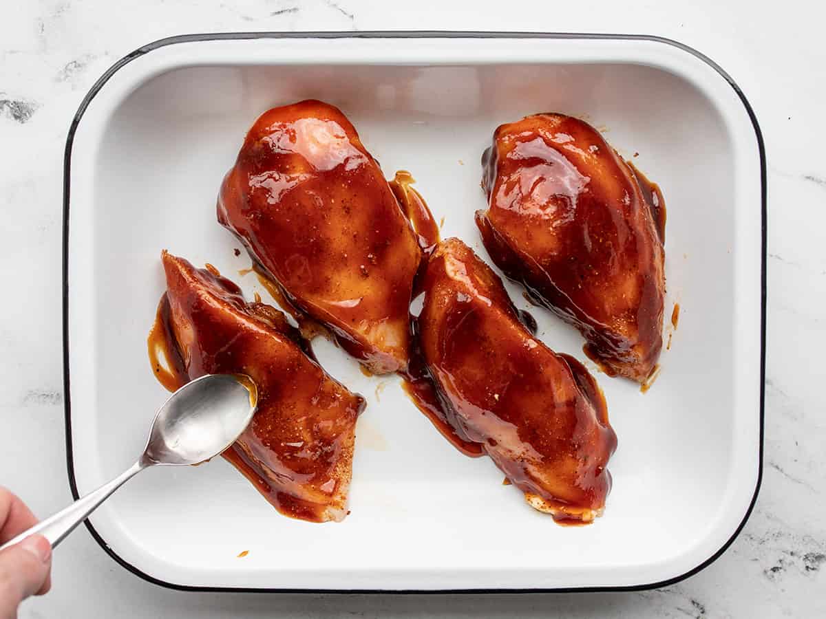 BBQ sauce being spread over chicken in a baking dish