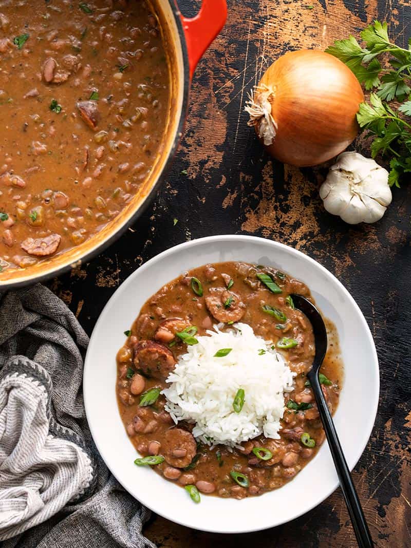 A serving of red beans and rice next to the pot full of red beans with an onion and garlic on the side.