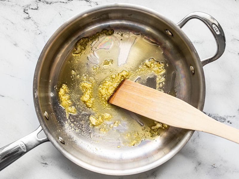 Minced garlic and grated ginger being sautéed in a skillet with a wooden spatula