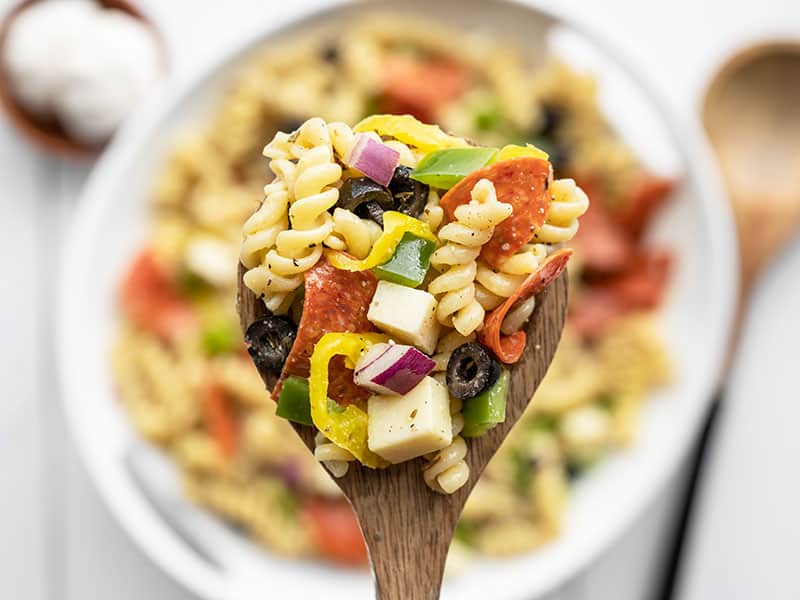 Close up of a large spoonful of pizza pasta salad on a wooden spoon, bowl in the background