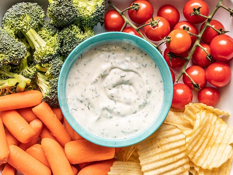A bowl of homemade ranch dip in the middle of broccoli, tomatoes, carrots, and potato chips