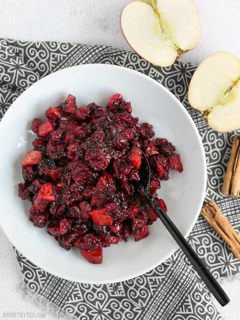 Roasted Apple Cranberry Relish in a white bowl with a black fork, on a patterned napkin, apples and cinnamon sticks on the side.