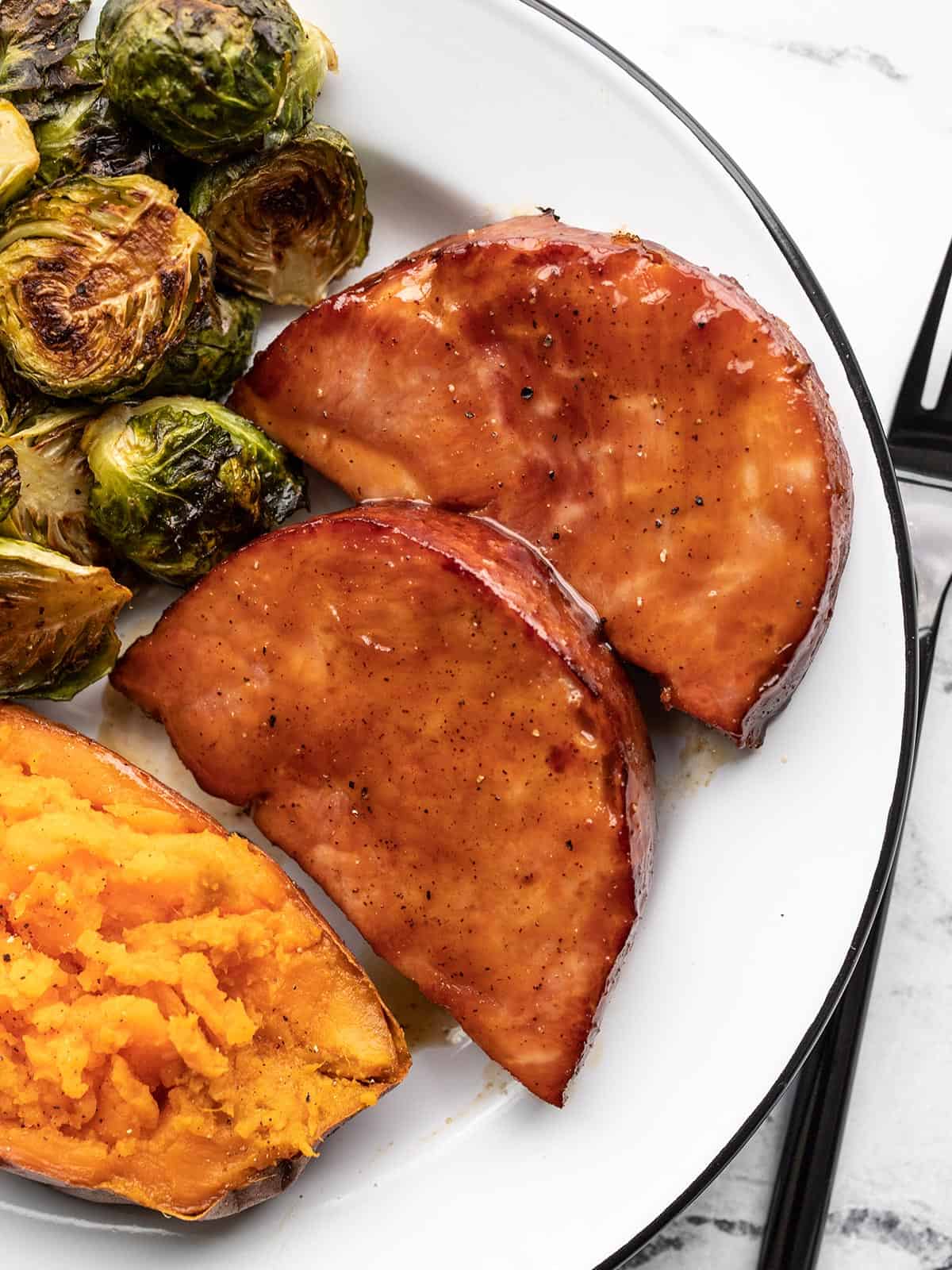 Two glazed ham steaks on a plate with Brussels sprouts and sweet potato