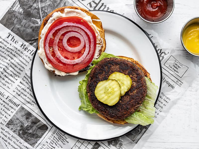 A black bean burger on a plate, dressed but open faced with ketchup and mustard on the side
