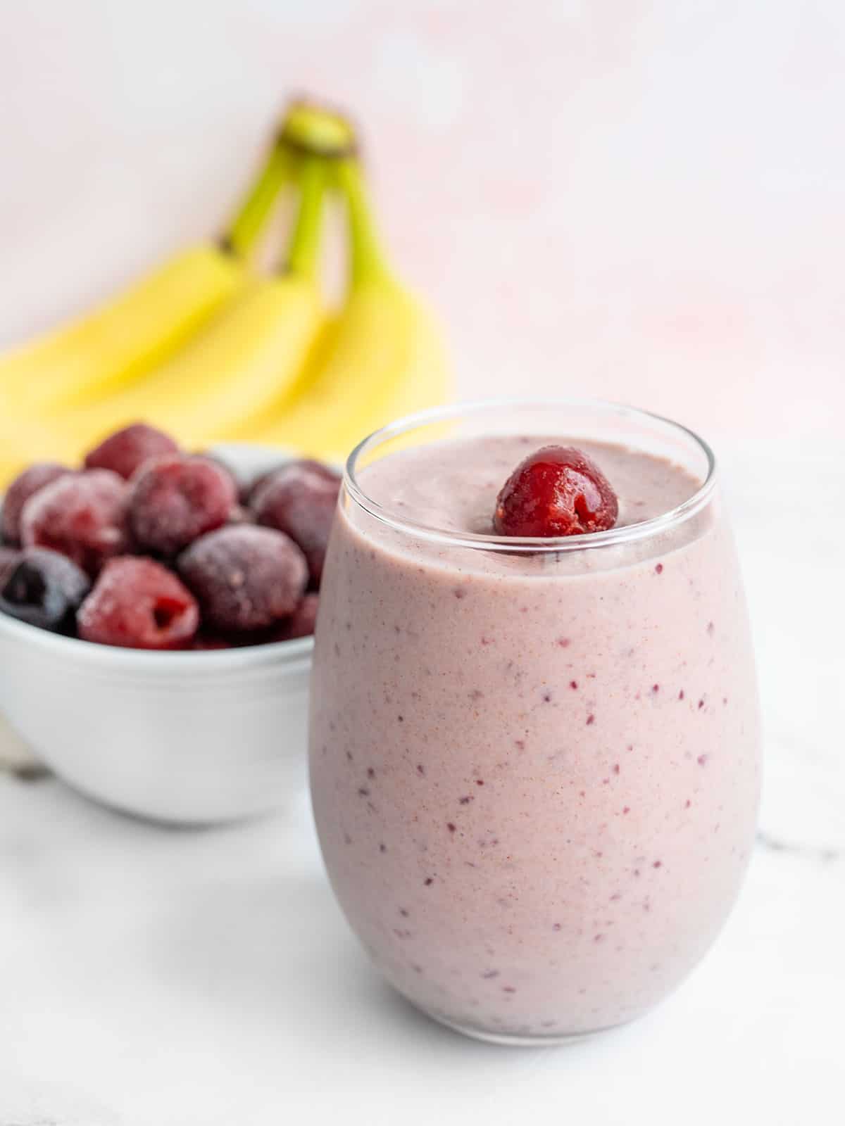 Cherry almond smoothie in a short glass with bananas and a bowl of frozen cherries behind it