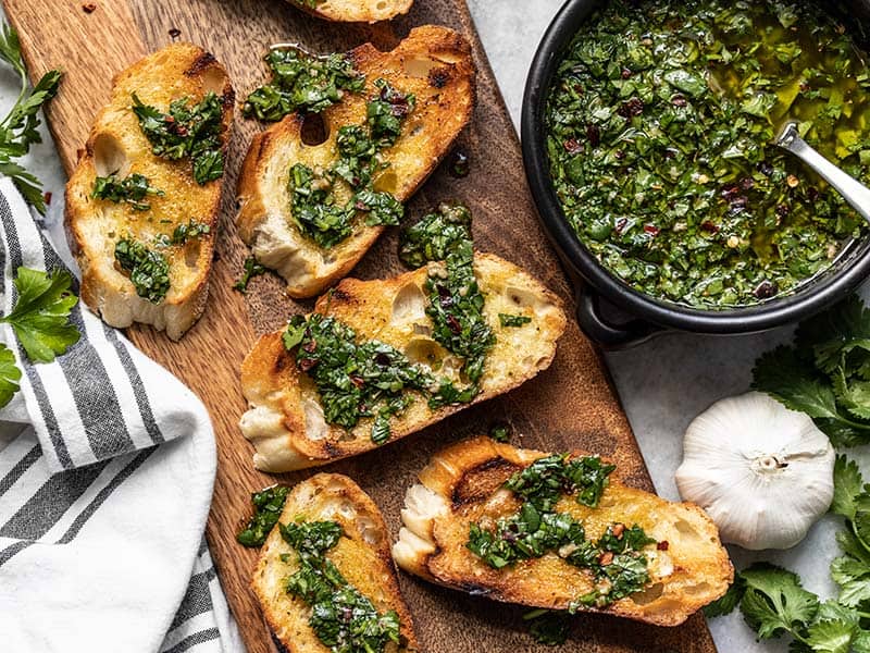 Sliced toasted French bread on a cutting board drizzled with chimichurri with a bowl of chimichurri sauce on the side.