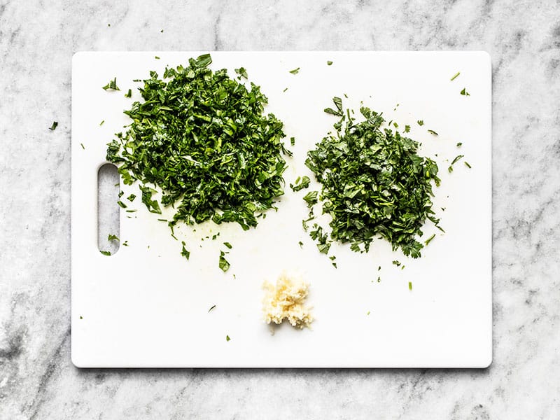 Chopped parsley and cilantro on a cutting board with minced garlic.