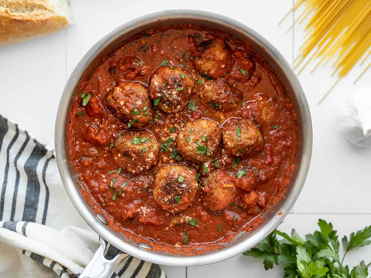 Meatballs in a pot of red sauce with spaghetti and herbs on the sides