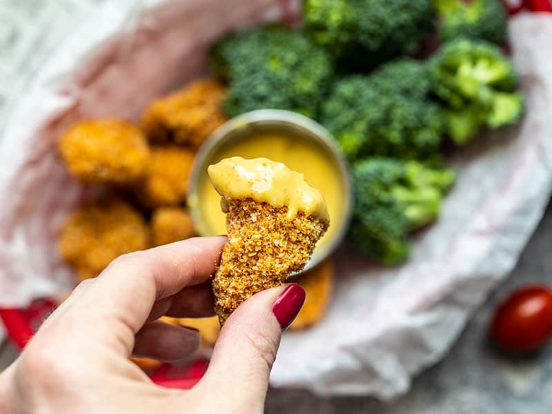 Close up of one Homemade Baked Chicken Nugget dipped in honey mustard with the basket of nuggets and vegetables in the background. 
