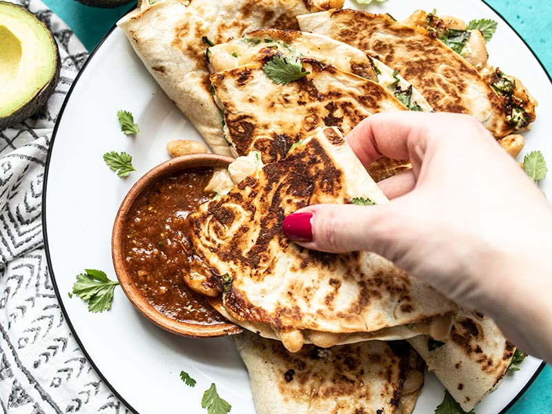 A hand dipping a piece of creamy white bean and spinach quesadilla into a bowl of red salsa