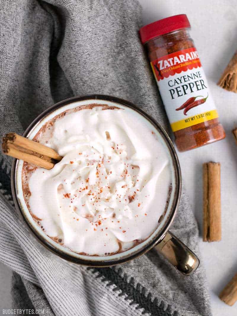 Overhead view of a mug full of Spicy Hot Cocoa topped with whipped cream and garnished with a cinnamon stick. A bottle of cayenne pepper on the side.