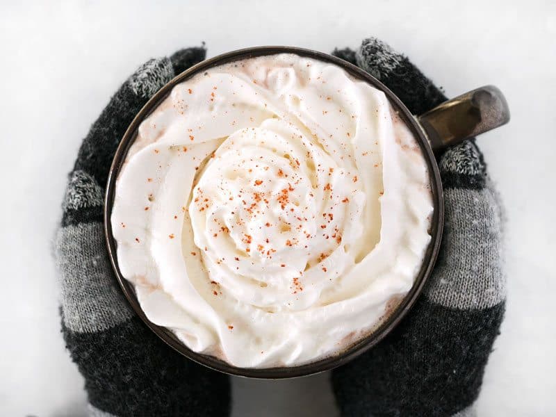 Overhead view of a pair of hands with gloves holding a mug full of spicy hot cocoa topped with whipped cream and a pinch of cayenne