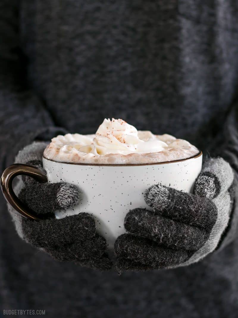 front view of a pair of hands with gloves holding a mug full of Spicy Aztec Hot Cocoa with whipped cream