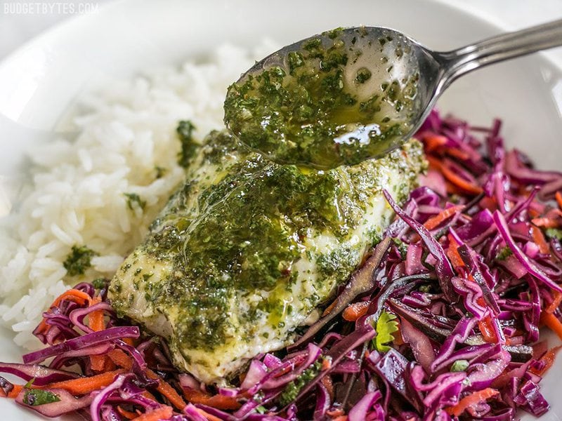 Chimichurri being drizzled over a piece of baked fish sitting on top of red cabbage slaw