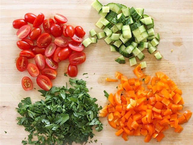 Chopped tomatoes, cucumber, bell pepper, and parsley on a wooden cutting board